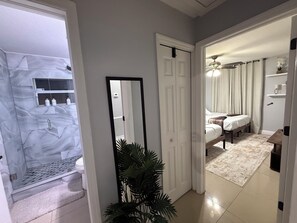 Corredor que leva a um quarto com cama, mesa de cabeceira e criado-mudo. Há um espelho na parede e uma planta no canto.