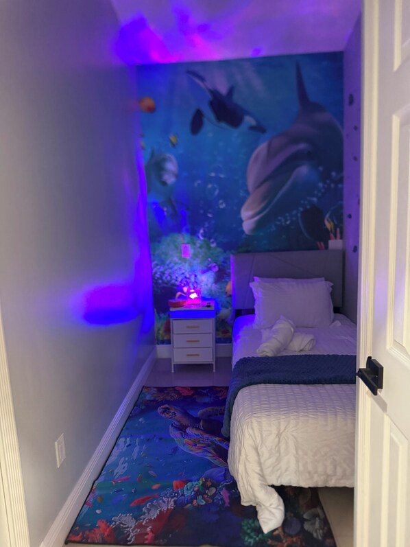 Een slaapkamer met een bed, een nachtkastje, een muurschildering en een kleurrijk vloerkleed.