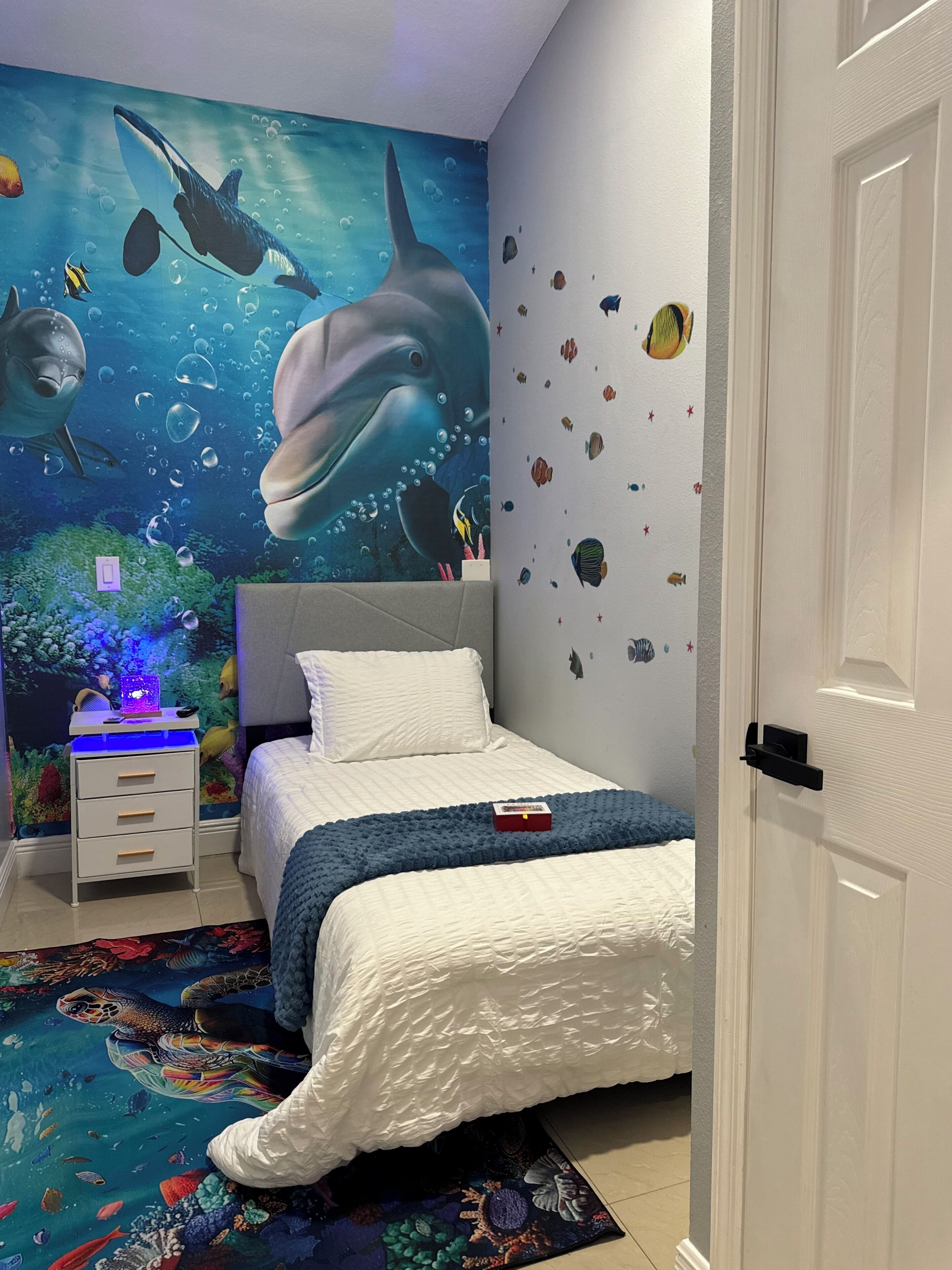 A bedroom with a mural of marine life, a bed with a blue blanket, a nightstand with a lamp, and a door.