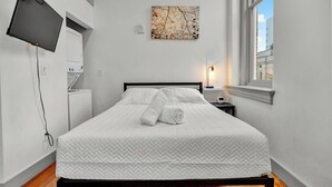 Une chambre d’hôtel comprenant un lit, un téléviseur à écran plat fixé au mur, un tableau au-dessus du lit, une lampe de chevet et une fenêtre donnant sur des immeubles.