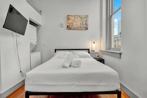 A hotel room with a bed, a flat-screen TV mounted on the wall, a painting above the bed, a bedside lamp, and a window with a view of buildings. - Executive Studio - Nearby Parking - Downtown - 3F (Louisville)