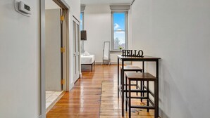 Un couloir avec un plancher en bois, une porte blanche, un panneau indiquant « BONJOUR » posé sur une table et une fenêtre donnant sur le ciel.