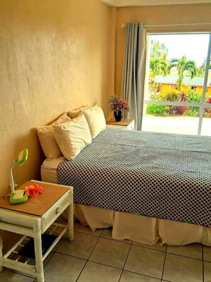 Ein Zimmer mit einem Bett, einem kleinen Tisch mit einer Lampe, einer Topfpflanze und Blick auf Grün.