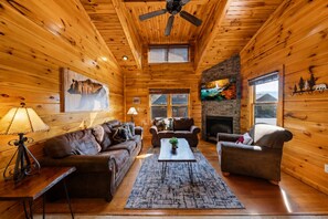 A log cabin interior with a fireplace, wooden walls, a ceiling fan, and a large window. - Sleeps 8, Mountain View Hot Tub, Arcade, Pool Table, 2 Mi to Gatlinburg Parkway (Gatlinburg)