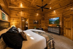 A log cabin bedroom with a bed, a ceiling fan, a fireplace, and a painting on the wall. - Sleeps 8, Mountain View Hot Tub, Arcade, Pool Table, 2 Mi to Gatlinburg Parkway (Gatlinburg)
