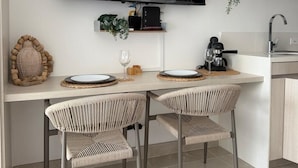 Uma cozinha moderna com uma televisão de tela plana montada acima de uma mesa de jantar, duas cadeiras de vime e uma cafeteira.