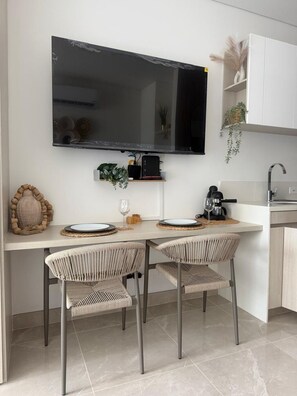 A modern kitchen with a flat-screen TV mounted above a dining table, two wicker chairs, and a coffee machine. - Apartment near the ocean at Bello Horizonte 1416 (Santa Marta)