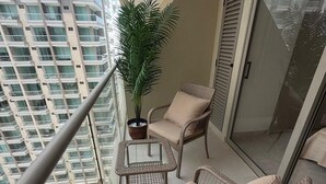 Uma varanda com móveis de vime, uma planta em vaso e vista para uma piscina e edifícios altos.