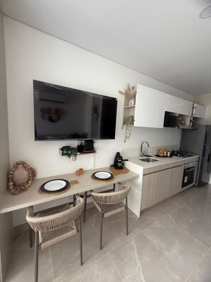 A modern kitchen with a flat-screen TV, white cabinets, a built-in oven, and a dining area with wicker chairs. - Oceanfront Apartment in Bello Horizonte 1416 (Santa Marta)