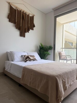 A bedroom with a bed, a macrame wall hanging, a potted plant, and a view of a balcony. - Oceanfront Apartment in Bello Horizonte 1416 (Santa Marta)