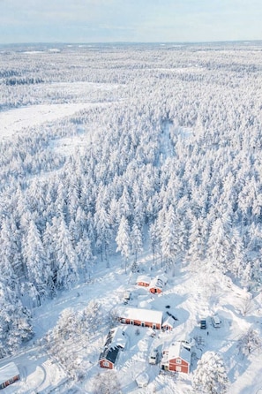 Un paisaje nevado con un grupo de edificios rojos, posiblemente una cabaña o alojamientos, rodeado de un bosque denso.