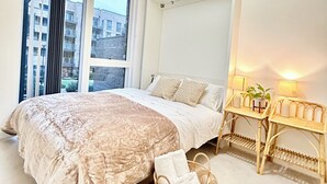 Et soveværelse med en stor seng, et natbord med lamper, en fletstol og udsigt til bygninger gennem vinduet.