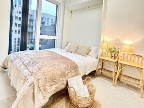 Ein Schlafzimmer mit einem großen Bett, einem Nachttisch mit Lampen, einem Korbstuhl und Blick auf Gebäude durch das Fenster.