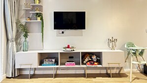 En moderne stue med et fladskærms-tv monteret på væggen, et hvidt tv-møbel med hylder, et lille bord og en høj stol.