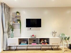 Ein modernes Wohnzimmer mit einem wandmontierten Flachbildfernseher, einer weißen Unterhaltungsecke mit Regalen, einem kleinen Tisch und einem Hochstuhl.