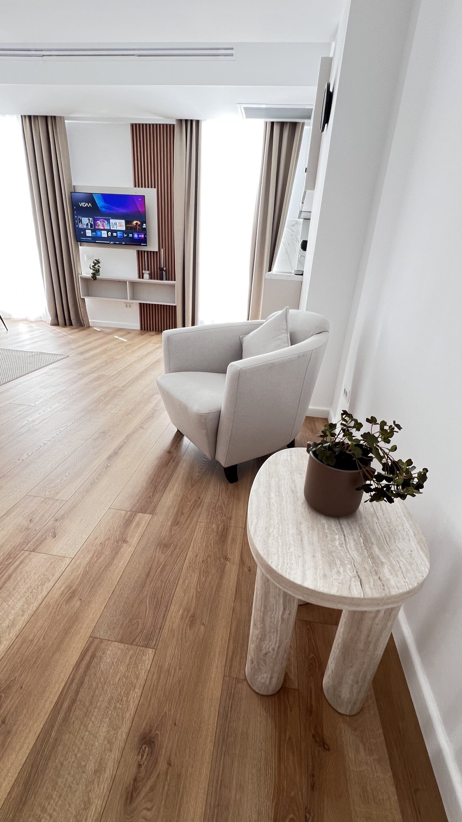 A modern living room with a flat-screen TV, a grey armchair, a round wooden side table with a plant, and a wooden floor.