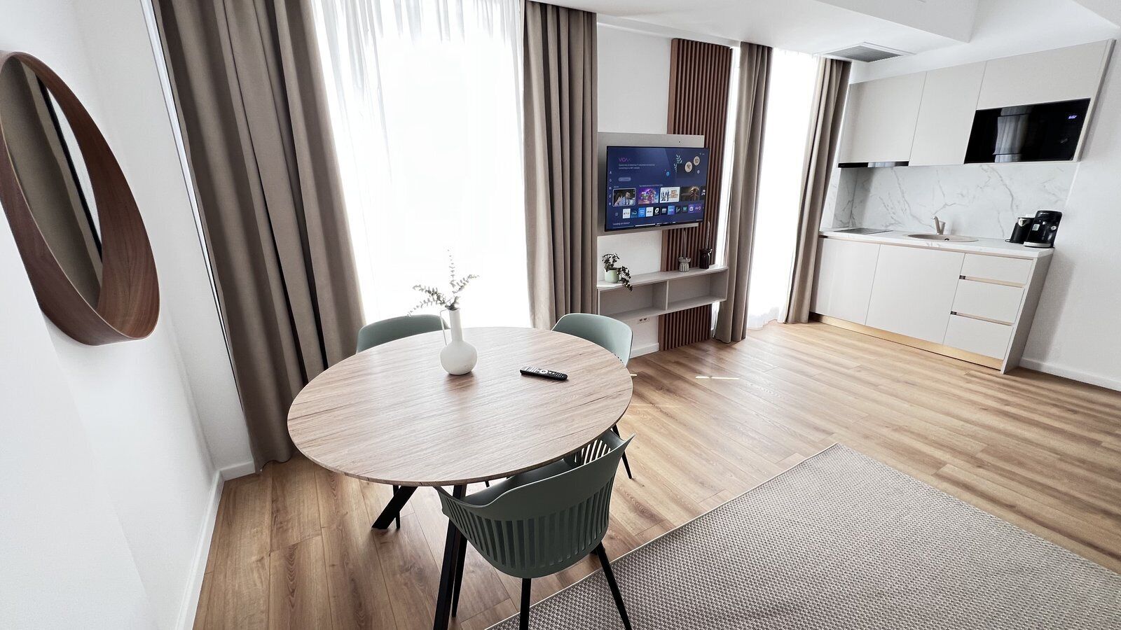 A modern hotel room with a round dining table, a TV mounted on the wall, a kitchen area, and a large window with curtains.