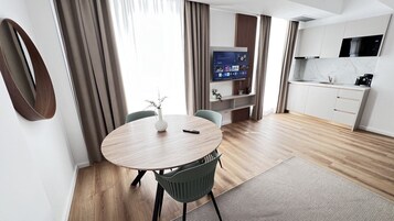 Moderni hotellihuone, jossa on pyöreä ruokapöytä, seinälle asennettu televisio, keittotila ja suuri ikkuna, jossa on verhot.