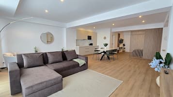 Ein modernes Wohnzimmer mit einem Ecksofa, einem Esstisch mit Stühlen, einer Küchenzeile und einem großen Spiegel an der Wand.