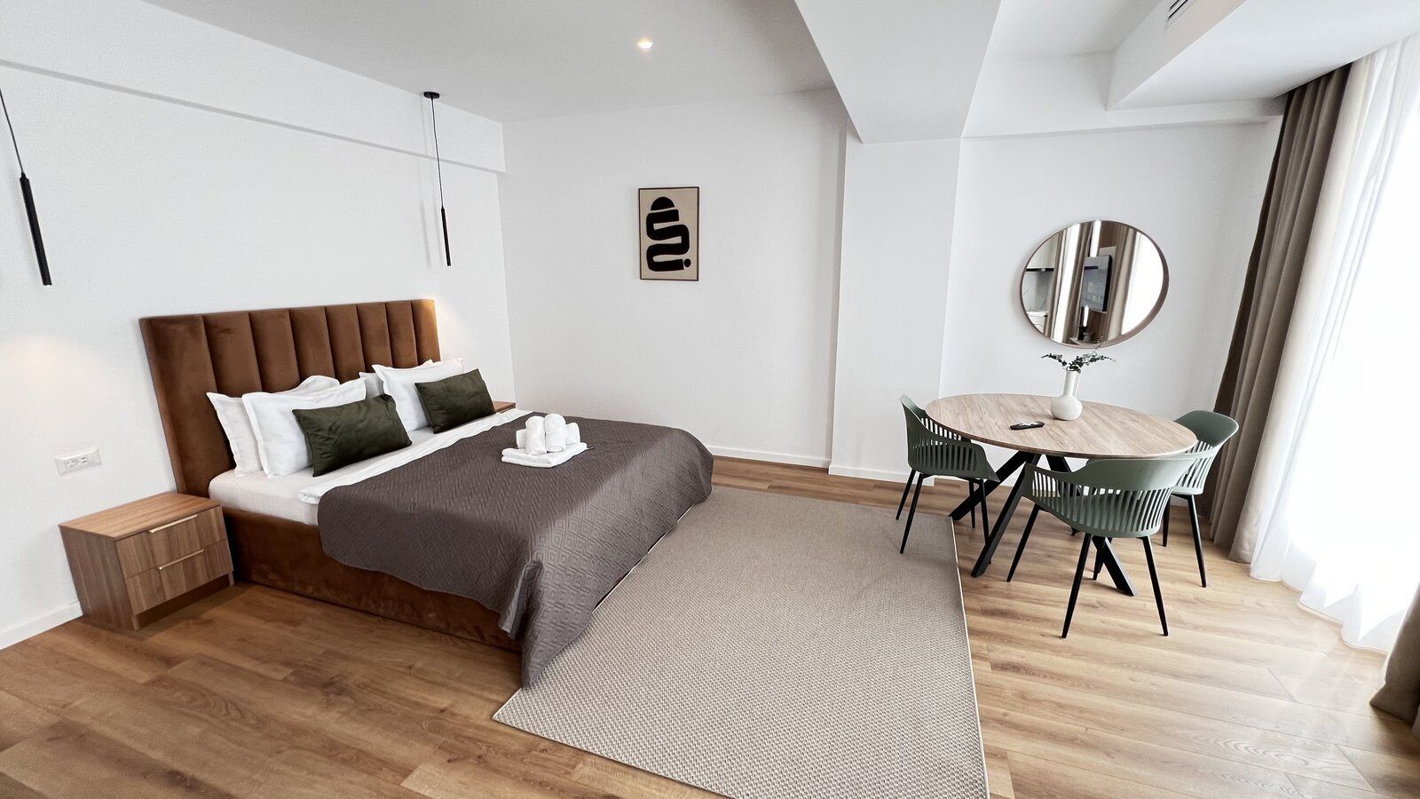 A modern hotel room with a bed, bedside table, a round dining table with chairs, a mirror, and a wall-mounted artwork.