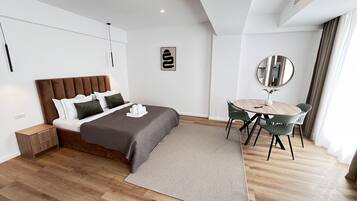 A modern hotel room with a bed, bedside table, a round dining table with chairs, a mirror, and a wall-mounted artwork.