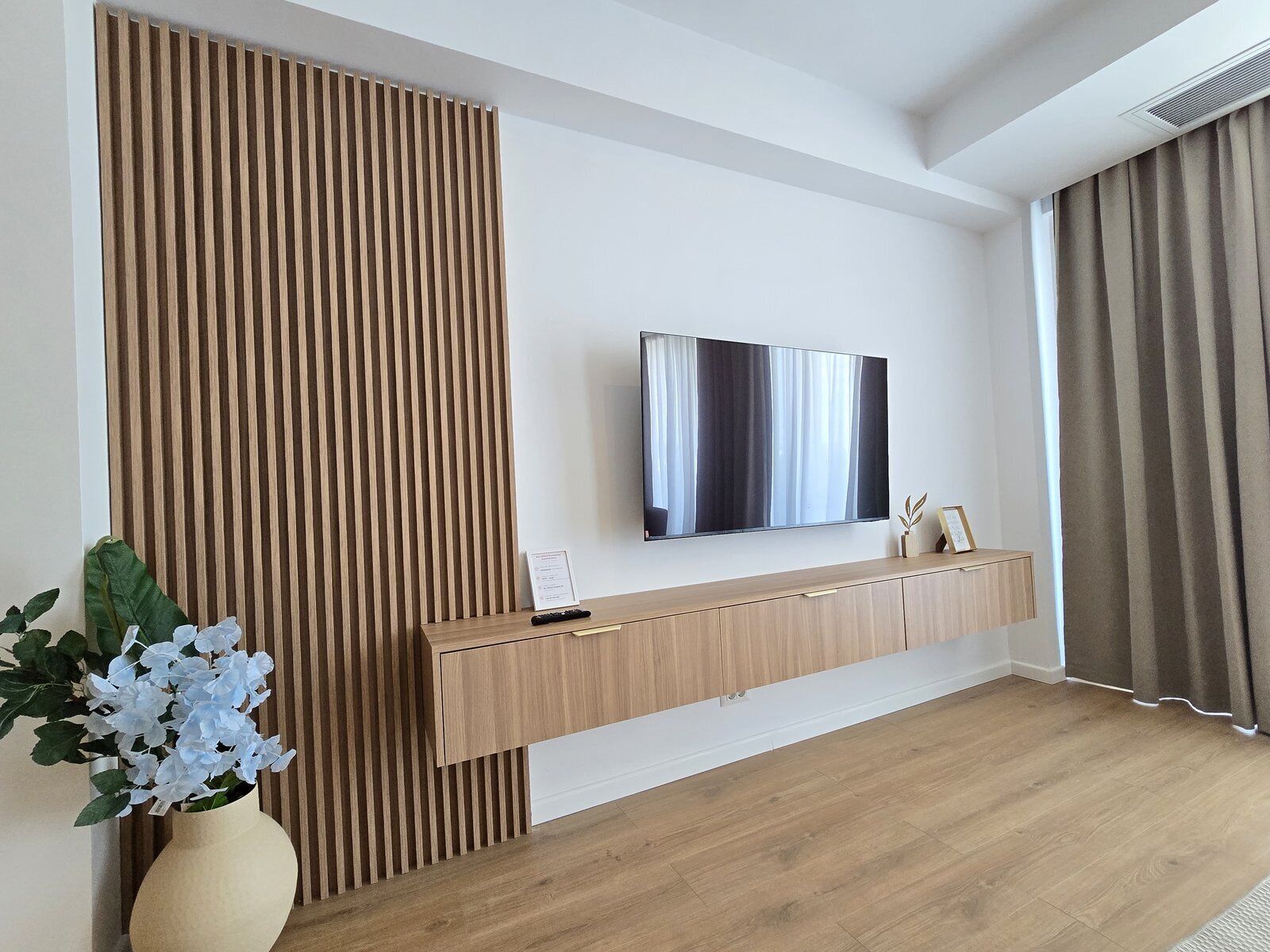 A modern living room with a wooden panel wall, a mounted TV, a console table, and a vase with flowers.