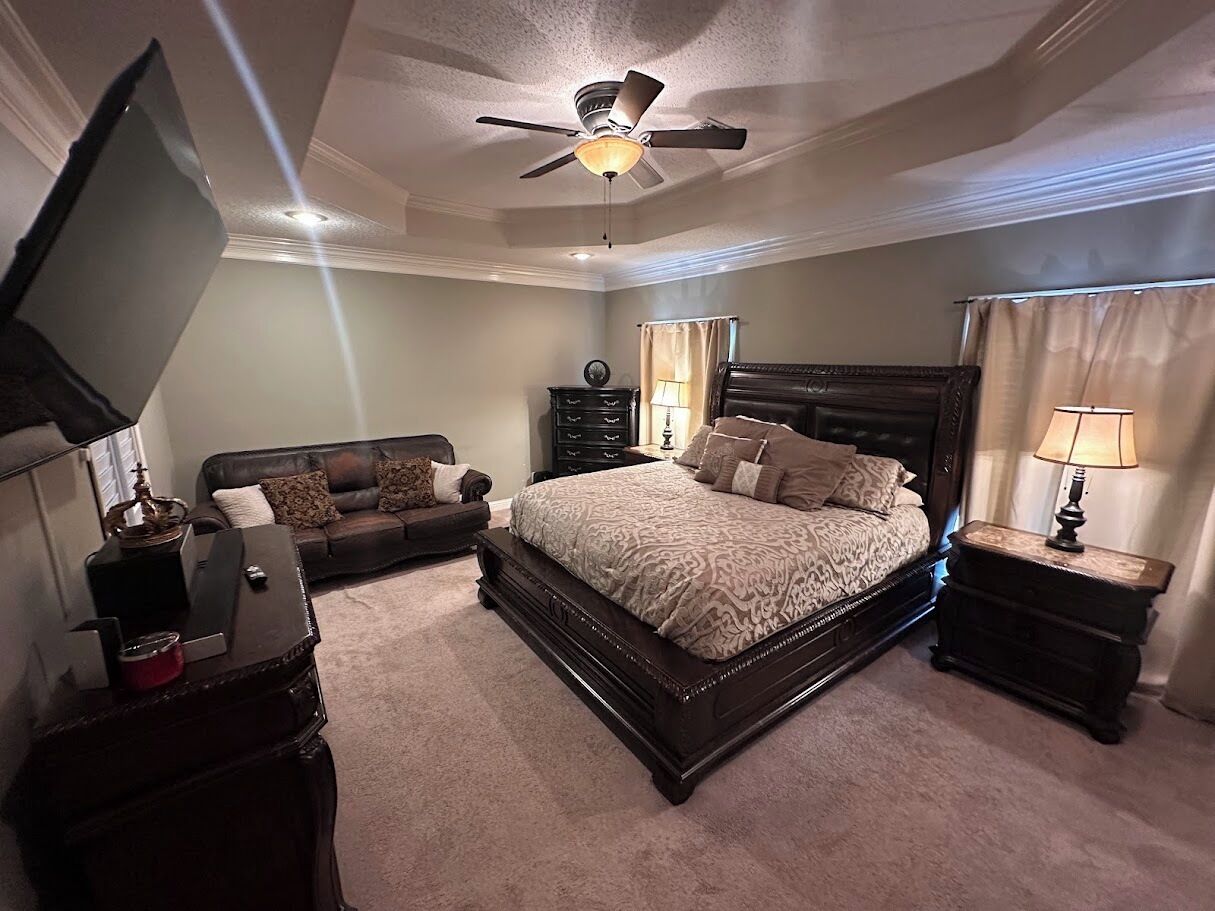 A bedroom with a large bed, a ceiling fan, a dresser, a nightstand, and a TV on a stand.