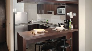 Una cocina moderna con alacenas de madera oscura, un refrigerador de acero inoxidable, un microondas y una cafetera. Hay dos taburetes de bar y una mesa de comedor con un plato y una taza blancos.