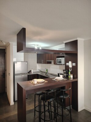 A modern kitchen with dark wood cabinets, a stainless steel refrigerator, a microwave, and a coffee maker. There are two bar stools and a dining table with a white plate and cup. - Oceana Suites Neohaus (La Serena)