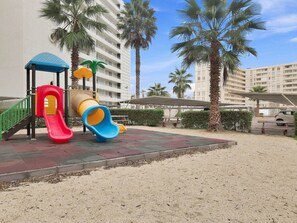A playground with slides and a climbing structure, surrounded by palm trees and a building in the background. - Oceana Suites Neohaus (La Serena)