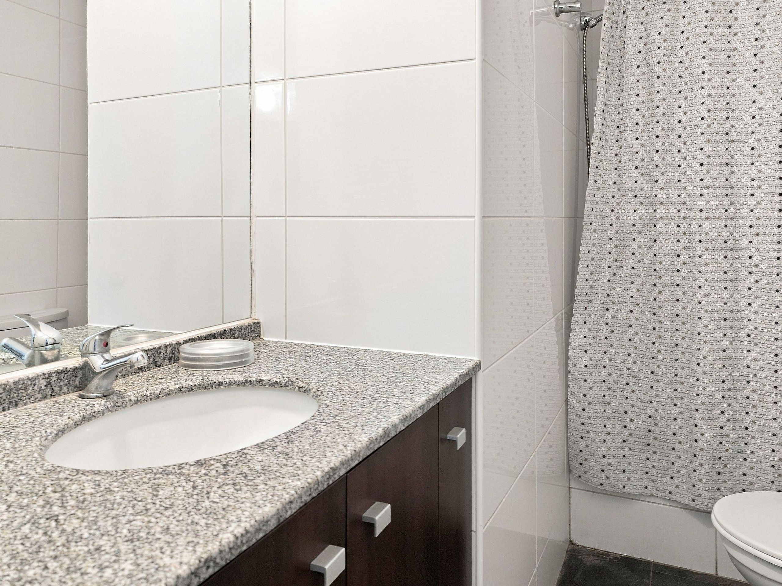 Un baño con lavamanos de encimera de granito, un espejo, cortina de ducha con estampado de lunares y un inodoro.