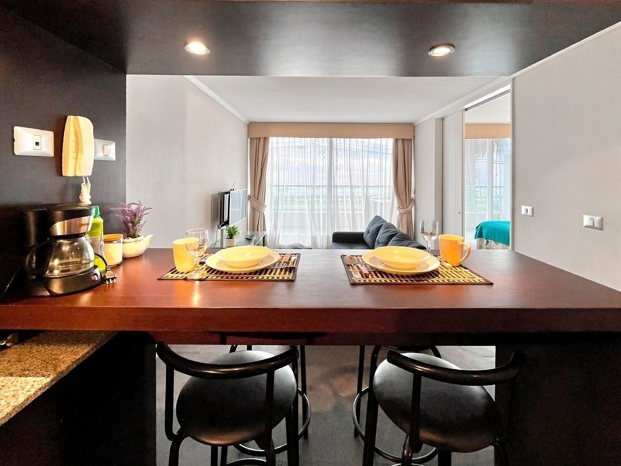 A modern kitchen with a dining table set for two, equipped with a coffee maker, a kettle, and a fruit bowl.