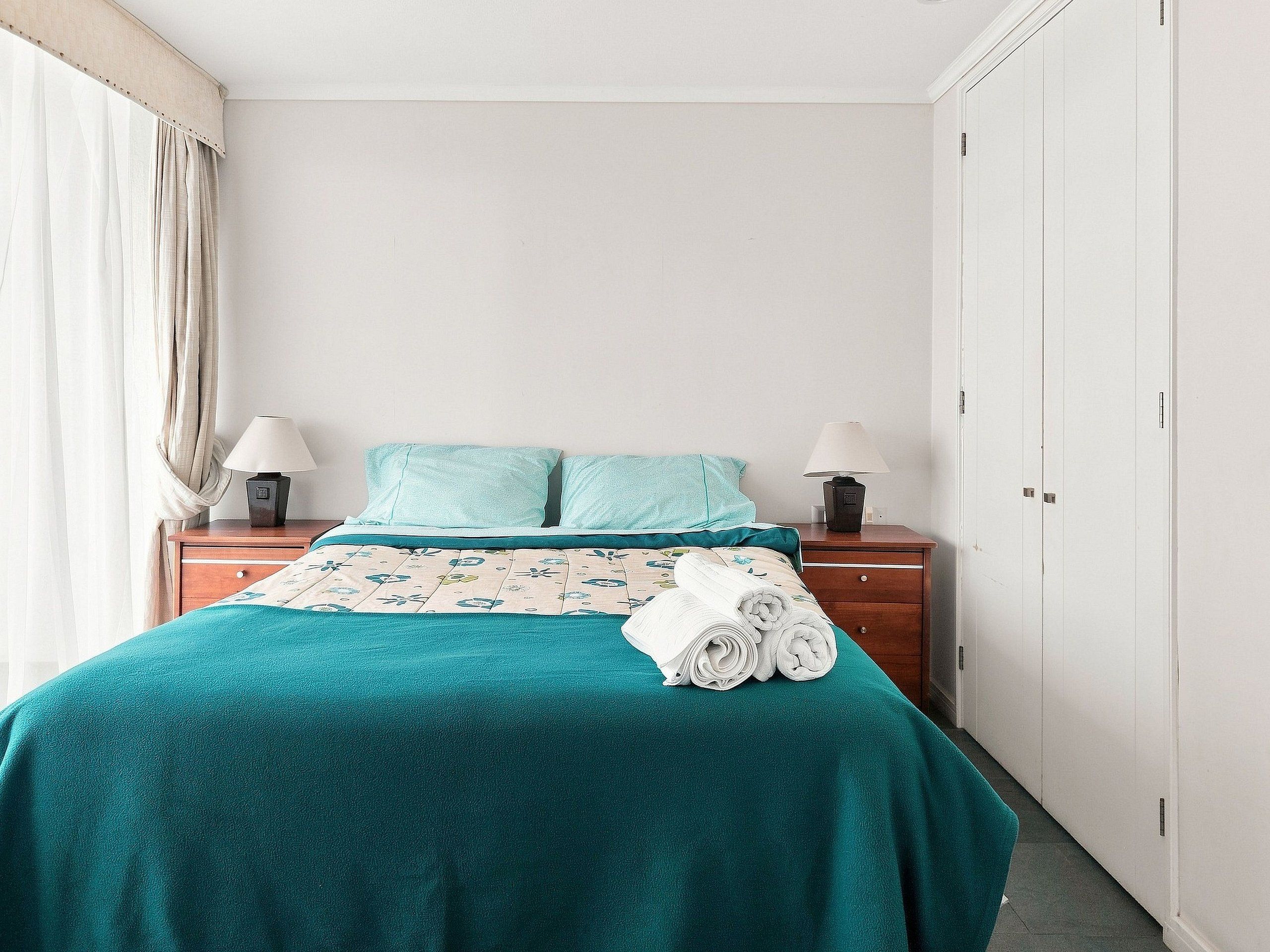 A hotel room with a bed, two bedside tables, a lamp on each table, a rolled towel on the bed, and a wardrobe.