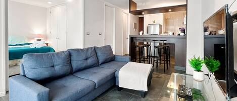 Un salon moderne avec un canapé bleu, une table basse en verre et un coin cuisine à l’arrière-plan.