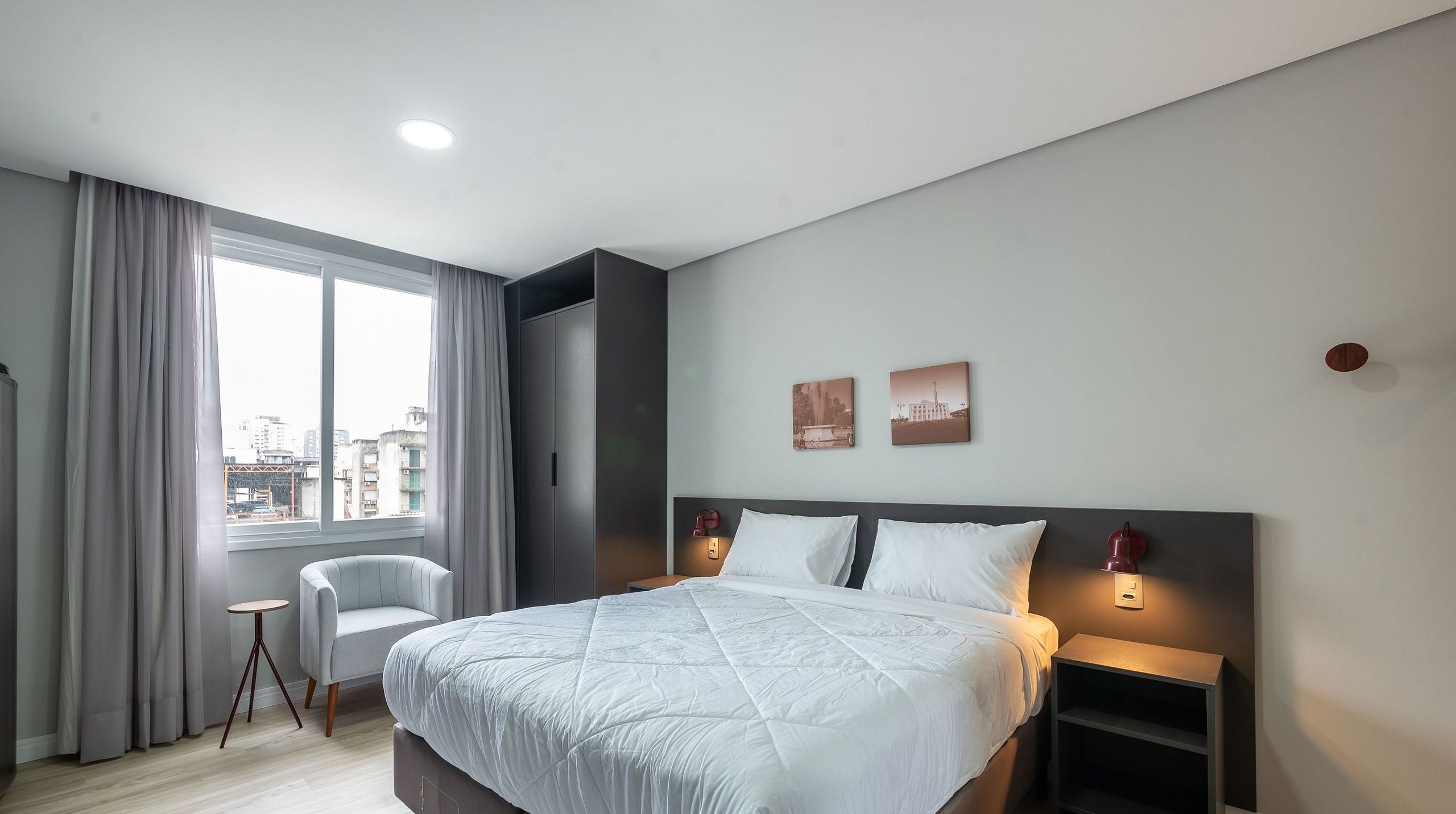 Quarto de hotel moderno com uma cama grande, uma cadeira, uma escrivaninha e vista para a cidade.