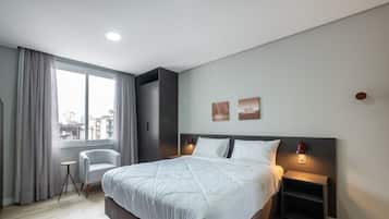 Quarto de hotel moderno com uma cama grande, uma cadeira, uma escrivaninha e vista para a cidade.