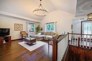 A spacious living room with a wooden floor, a large L-shaped sectional sofa, a coffee table, a television on a wooden stand, and a chandelier.