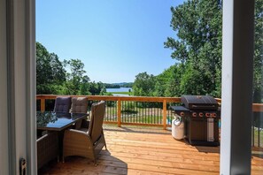 Una terraza de madera con mobiliario exterior, una parrilla y vista a un lago.