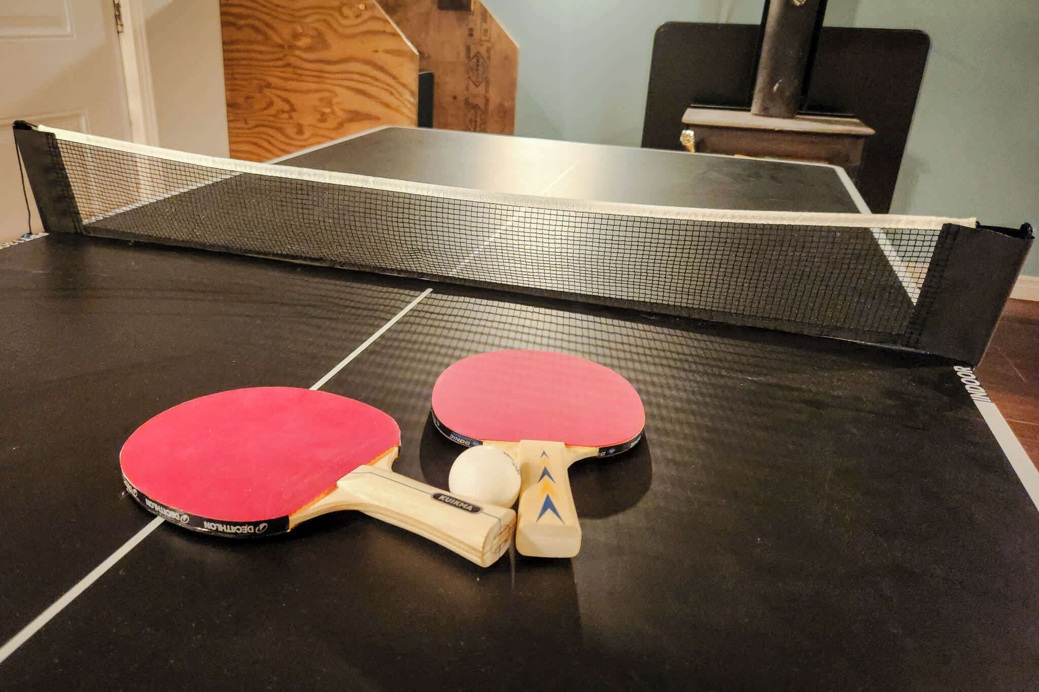 A ping pong table with two red paddles and a ball, set in a room with a wooden floor and a black table.