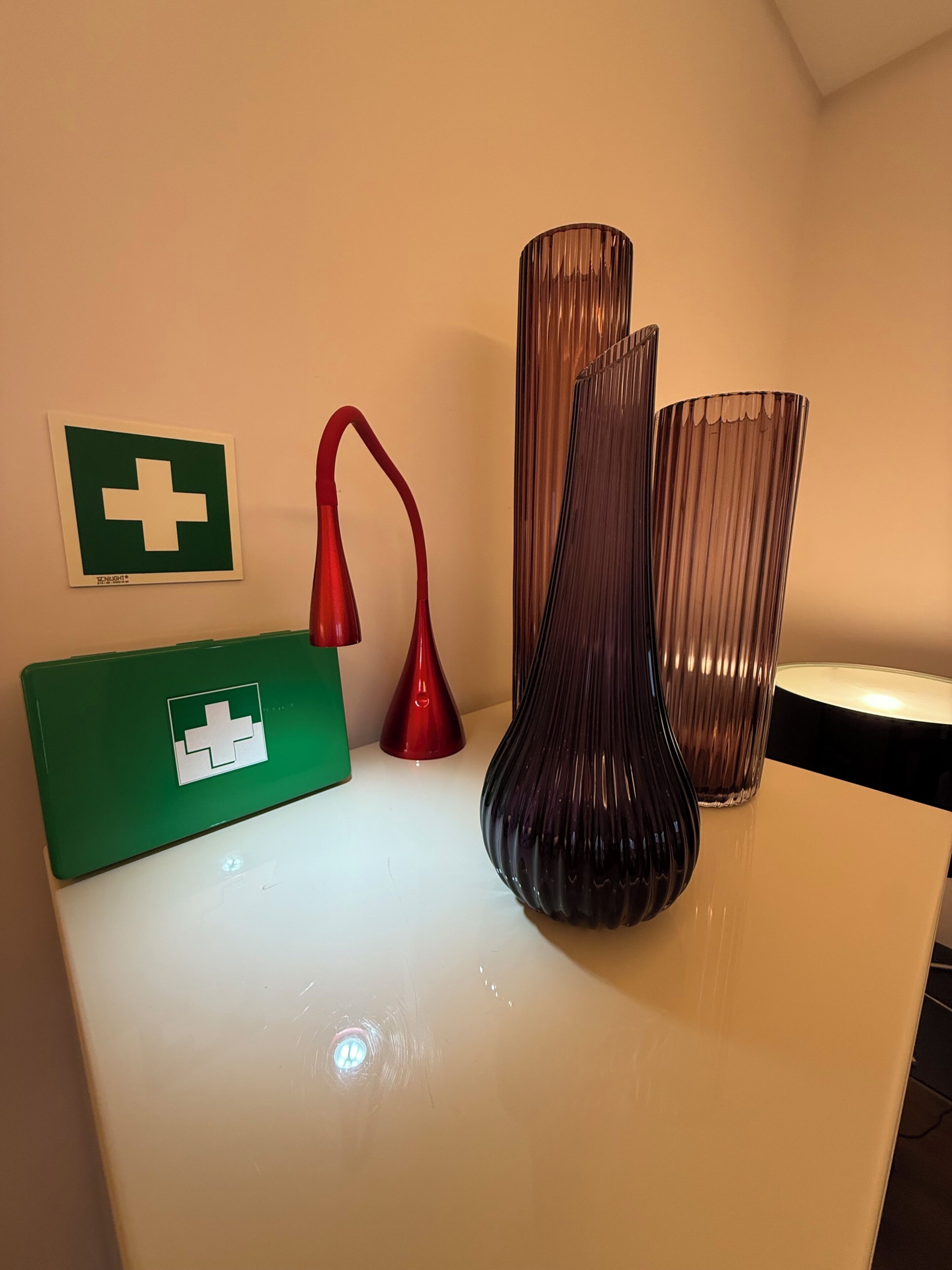 A white table with three vases, a red lamp, and a green box with a white cross symbol.
