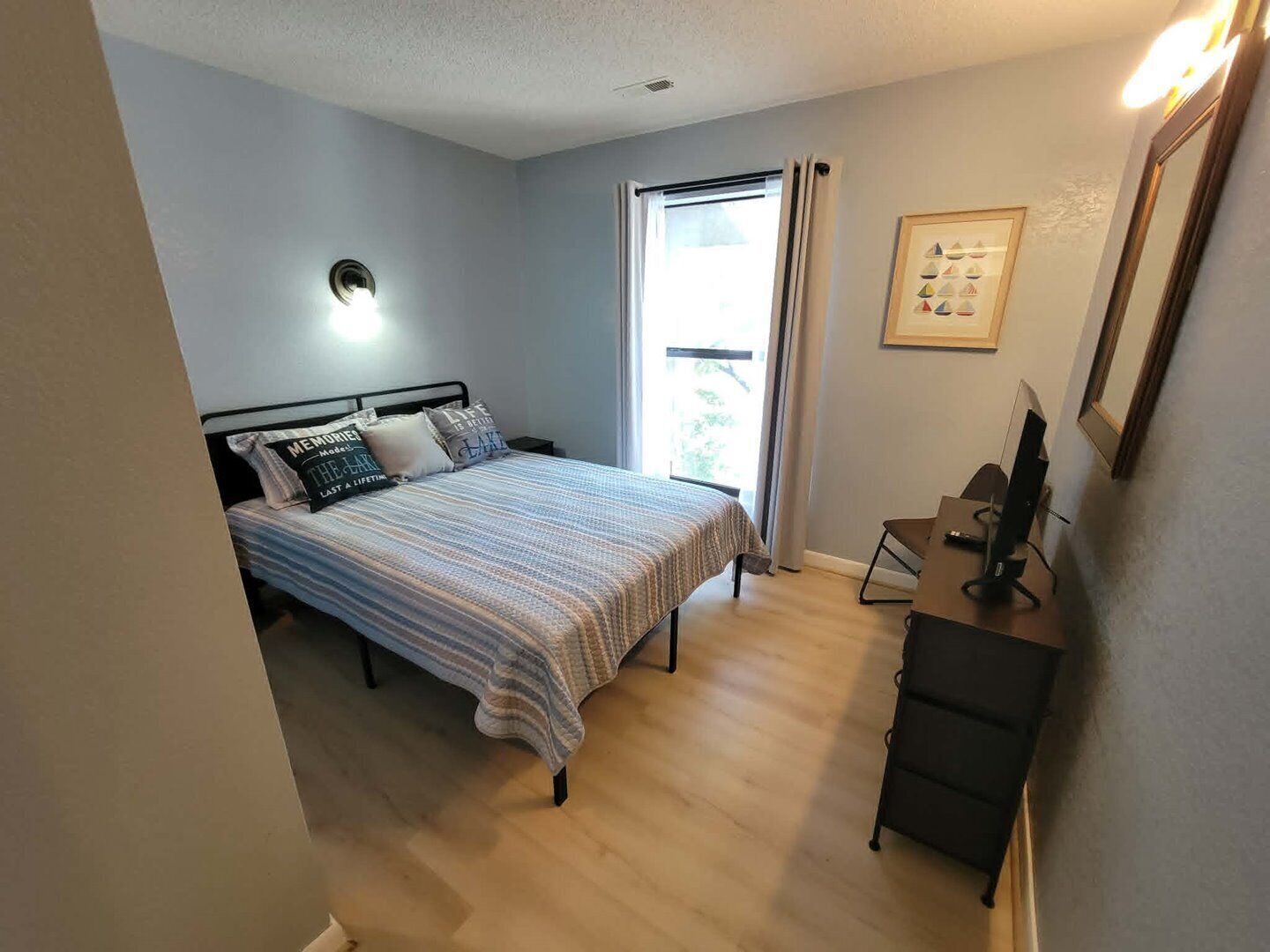 A bedroom with a bed, a desk, a chair, a window, and a wall-mounted light fixture.