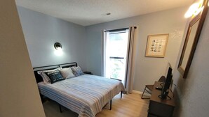 Ein Schlafzimmer mit Bett, Schreibtisch, Stuhl, Fenster und einer Wandleuchte.