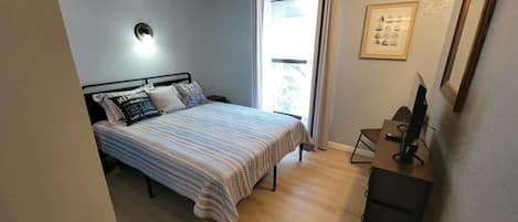 Un dormitorio con cama, escritorio, silla, ventana y una lámpara de pared.