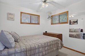 Ein Schlafzimmer mit einem Bett, einer Kommode, zwei Fenstern mit Jalousien, einem Deckenventilator und einem an der Wand befestigten Spiegel.