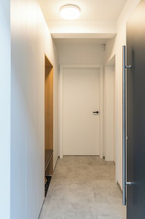 A narrow hallway with a white door at the end, a wooden bench on the left, and a dark-colored refrigerator on the right. - Peaceful 2-bedroom apartment in Bled's remote area perfect for relaxing (Bled)