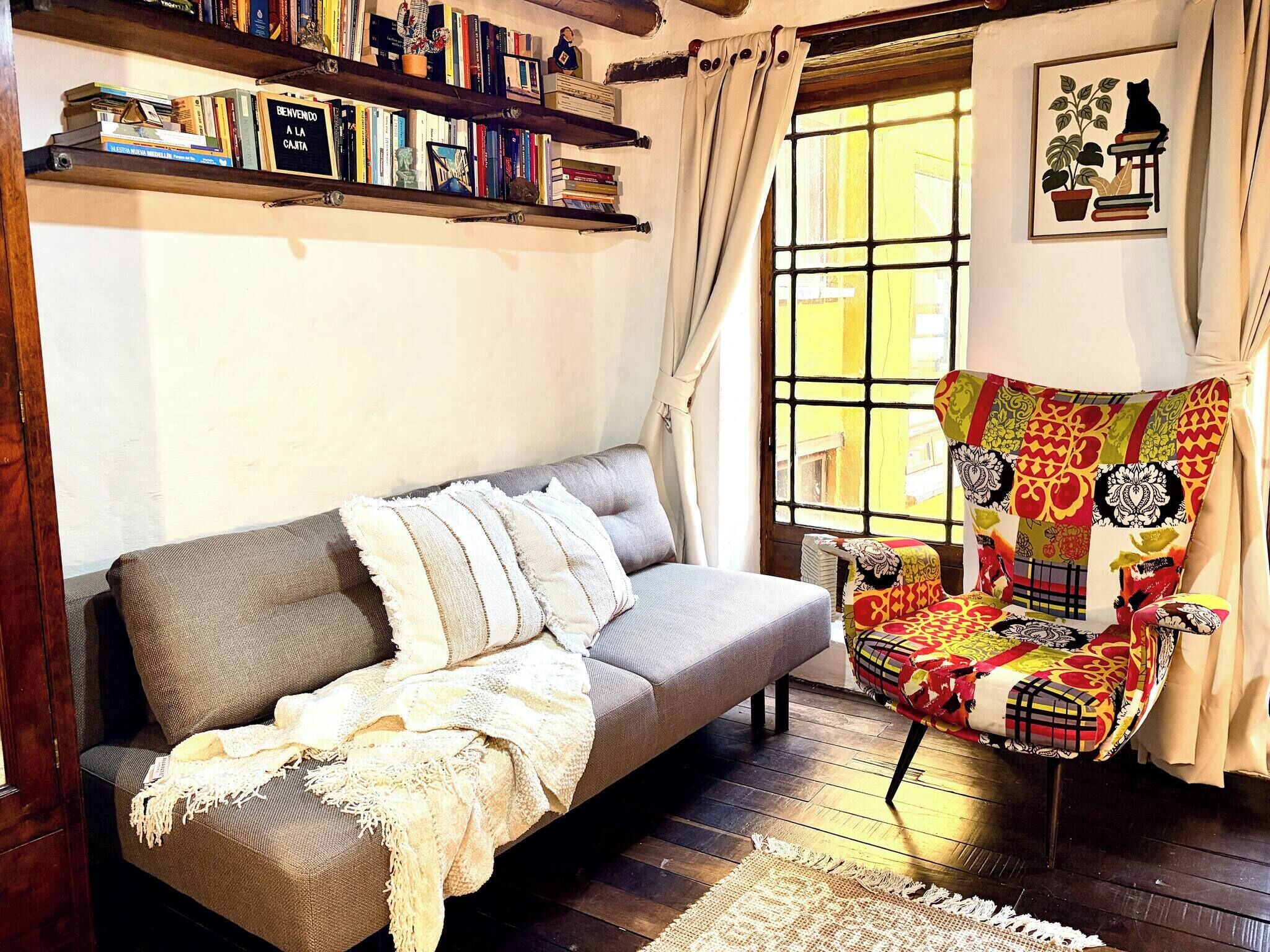 A cozy living room with a sofa, a colorful armchair, a bookshelf, and a window with curtains.