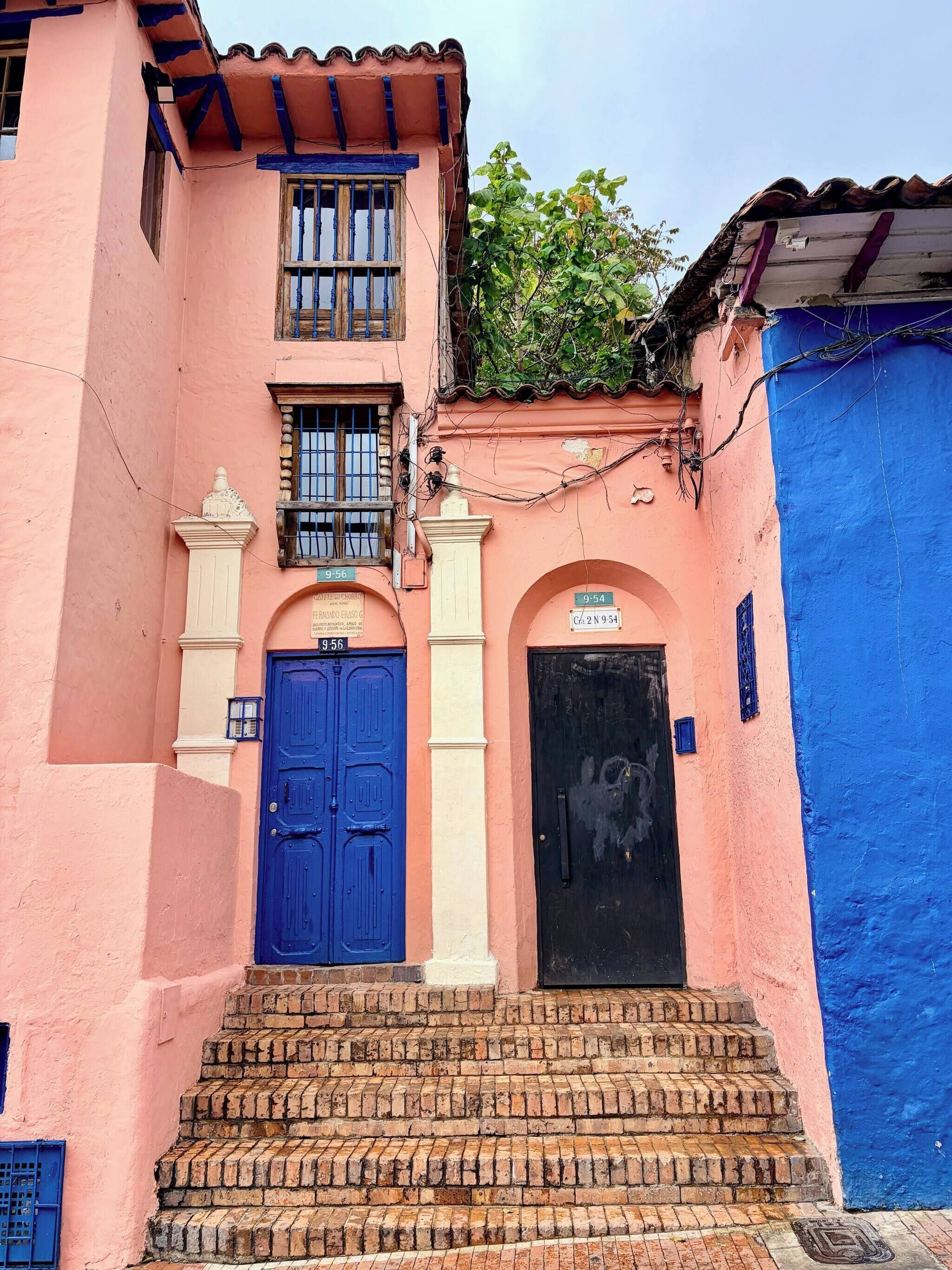 A pink building with a blue door and a black door, a staircase, and a small balcony with a plant.
