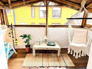 A cozy room with a hammock, a small table with decorative items, a potted plant, and a view of a yellow building. - Iechie Historic Colonial Stay in La Candelaria (Bogotá)