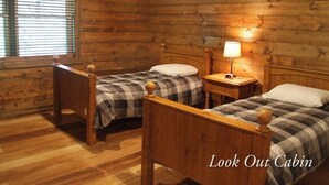 A cabin-style room with two wooden beds, a nightstand with a lamp, and a window with blinds. - Lookout Cabin 1 (Caledonia)
