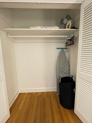Ein begehbarer Kleiderschrank mit Holzboden, einer Kleiderstange voller Kleidung, einem Ventilator und einer Flasche auf einem Regal.
